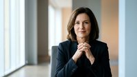 Middle-aged woman with dark hair and Caucasian ethnicity wearing professional black blazer, hands clasped together, confident expression in modern office corridor with natural lighting.