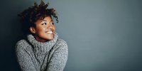 Young African American woman with curly hair smiling while wearing cozy gray turtleneck sweater against neutral background, conveying warmth and authenticity.