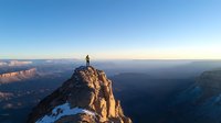 Lone hiker stands triumphantly on mountain peak at sunrise, overlooking vast canyon landscape with snow patches and dramatic cliffs in golden morning light.