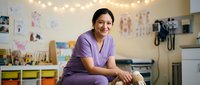 Female healthcare professional of South Asian descent wearing purple scrubs, smiling while posing in a modern medical clinic or pediatric office with colorful storage, educational posters, and warm string lights overhead.