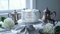 Elegant white birthday cake with silver stars on marble stand, surrounded by hydrangeas and vintage silver teapots on a soft gray table.