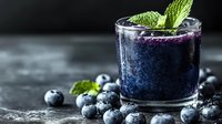 Fresh blueberry smoothie garnished with mint leaves in glass on dark background with scattered berries and condensation droplets. Perfect for menu design.