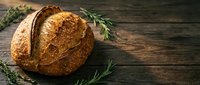 Freshly baked artisan sourdough bread loaf with golden crust on rustic wooden table with rosemary herbs for bakery and homemade cooking concepts.