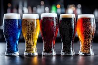 Five different colored beer glasses with fresh foam lined up on dark bar counter showing variety of craft brews from light to dark with bokeh background.