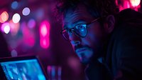 Young caucasian male programmer in glasses working late at night on laptop with neon pink and purple lighting creating dramatic cyberpunk atmosphere.