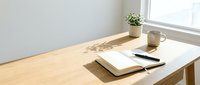 Minimalist workspace with open notebook, pen, and glass of water on wooden desk near bright window with natural light and potted plant.