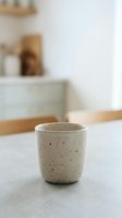 Minimalist ceramic cup with speckled glaze sitting on concrete countertop in bright, airy kitchen with blurred cabinetry and neutral tones for home decor and lifestyle use.