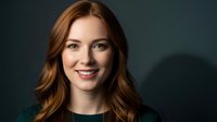 Professional young Caucasian businesswoman with wavy auburn hair smiling confidently against dark background for corporate headshot and marketing materials.