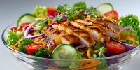 Grilled chicken breast sliced over fresh mixed greens with cherry tomatoes, cucumber and colorful vegetables in glass bowl for healthy dining.