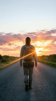 Woman jogging on rural road at golden hour sunset, viewed from behind. Long-haired female athlete in casual sportswear running toward vibrant orange sky.