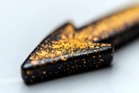 Macro shot of black arrow tip covered with golden glitter particles on white background, shallow depth of field creates dreamy bokeh effect.