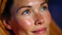 Close up portrait of young woman with freckles, glowing skin and bright blue eyes looking up dreamily against dark background, wearing orange headscarf.