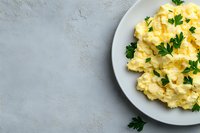 Creamy scrambled eggs garnished with fresh parsley on white plate against textured gray background for breakfast menu design and culinary content.