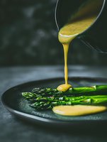 Pouring golden hollandaise sauce over fresh grilled asparagus spears on dark plate, dramatic food photography with moody lighting and selective focus.