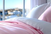 Cozy bedroom interior with soft pink and white bedding, pillows against window with blurred coastal view background, creating serene morning atmosphere.