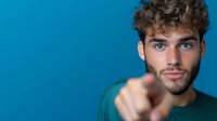 Young Caucasian man with curly hair and beard pointing finger at camera against blue background, expressing confidence and direct engagement in casual green shirt.
