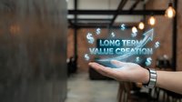 Long term value creation concept with upward arrow and dollar symbols displayed as holographic projection above open palm in modern office environment.