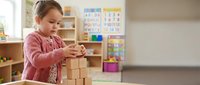 Young girl building tower with wooden blocks in bright classroom setting, developing fine motor skills and creativity through educational play activities.