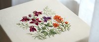 Embroidered wildflower bouquet on cream fabric featuring purple and orange blooms with green stems in traditional needlework style for crafting projects.