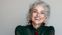 Elegant senior woman with silver gray hair wearing emerald green lace dress and red statement earrings, showing genuine happy smile against neutral background.