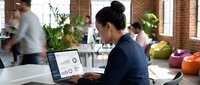Asian businesswoman analyzing financial data on laptop in modern coworking office space with colleagues working in background during busy workday.