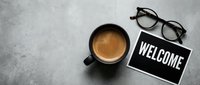 Black coffee mug, welcome sign chalkboard, and reading glasses on gray concrete surface, flat lay top view for business or hospitality concept.