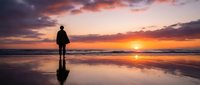 Silhouetted surfer holding board on wet beach at golden sunset, standing in shallow water with dramatic orange and purple sky reflecting on sand, perfect for travel and lifestyle content.