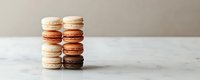 Stacked French macarons in various shades from white to brown on marble surface against neutral background, showcasing delicate meringue-based confections for bakery menu.