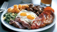 Hearty breakfast plate with sunny side up eggs, crispy bacon, ham, roasted potatoes, sauteed mushrooms and fresh broccoli served on white ceramic plate, close up view.