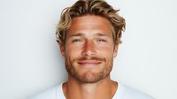 Confident young Caucasian man with blonde wavy hair and beard wearing white t-shirt smiling genuinely against light background, expressing positive attitude and friendliness.