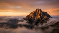 Majestic mountain peak emerging from clouds at golden hour sunrise with dramatic orange sky and misty atmosphere creating breathtaking alpine landscape scenery.