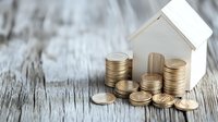 Small white house model with stacked gold coins on rustic wooden surface, representing real estate investment, mortgage, and property financial planning.