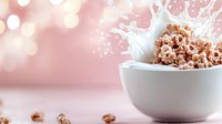 Crispy chocolate cereal rings splashing in white milk bowl against soft pink bokeh background, creating dynamic motion and appetizing breakfast scene.