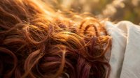 Young woman with flowing red curly hair in golden sunlight creating warm bokeh effect. Natural beauty and hair care concept for wellness lifestyle.