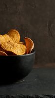 Crispy golden potato chips with seasoning in dark ceramic bowl on slate surface with moody lighting for food photography and culinary content.