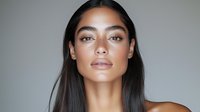 Young Hispanic woman with glowing natural makeup and straight dark hair against gray background, fresh dewy skin and neutral expression for beauty and skincare.