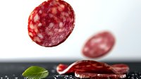 Sliced salami falling on black surface with green basil leaf, creating dynamic food composition for culinary photography.