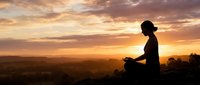 Woman meditating in lotus pose during golden hour sunset with dramatic sky and mountain landscape silhouette for wellness and mindfulness concepts.