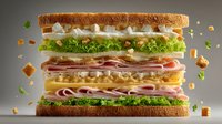 Towering deli sandwich with multiple layers of meats, cheese, lettuce and tomatoes on toasted bread with floating ingredients around it.