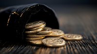 Golden coins spilling from black velvet pouch on rustic wooden surface, dramatic lighting creates contrast between shiny metal and dark background.