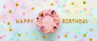 Pink birthday cake with floral decorations and golden Happy Birthday candles, surrounded by colorful confetti on a pastel background, top view.
