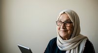 Mature woman with white hair, wearing glasses and a white headscarf over dark clothing, holding a tablet device while smiling at camera against neutral background for business, technology, and lifestyle use.
