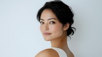 Young Asian woman with natural makeup and messy updo hairstyle wearing white top, turning and smiling against light background. Soft beauty portrait with clean skin.