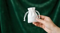 Female hand with medium skin tone holding white cotton drawstring pouch against deep green fabric background for product packaging, gift wrapping, retail display, and branding mockups.