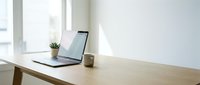 Laptop on wooden desk near window with small plant and coffee cup, bright natural light workspace for remote work and productivity.