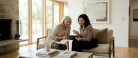 Senior woman with gray hair and younger woman with dark curly hair discussing documents together on sofa in bright modern living room for financial planning, consulting, or family meeting.