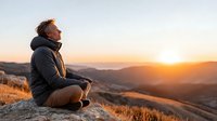 Middle-aged man in winter jacket sitting on mountain peak during golden hour sunrise, contemplating peaceful landscape with rolling hills and warm light.