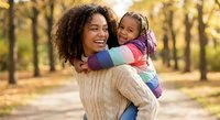 Happy African American mother holding smiling toddler daughter in autumn park with golden sunlight and colorful fall foliage creating warm family moment.
