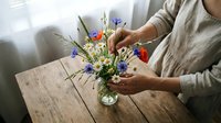 Woman arranging colorful wildflower bouquet in glass vase on rustic wooden table by window, creating natural home decor arrangement with fresh blooms.