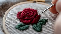 Detailed embroidery process showing red rose with green leaves on natural linen fabric in wooden hoop, close up view of needlework technique in progress.
